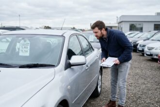 Homme examinant une voiture d'occasion dans un parking de concessionnaire avec liste de vérification à la main